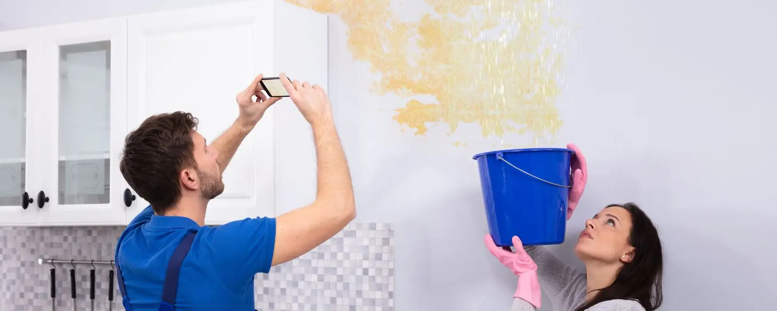 Man photographing a water-stained ceiling while a woman holds a bucket