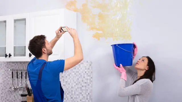 Man photographing a water-stained ceiling while a woman holds a bucket